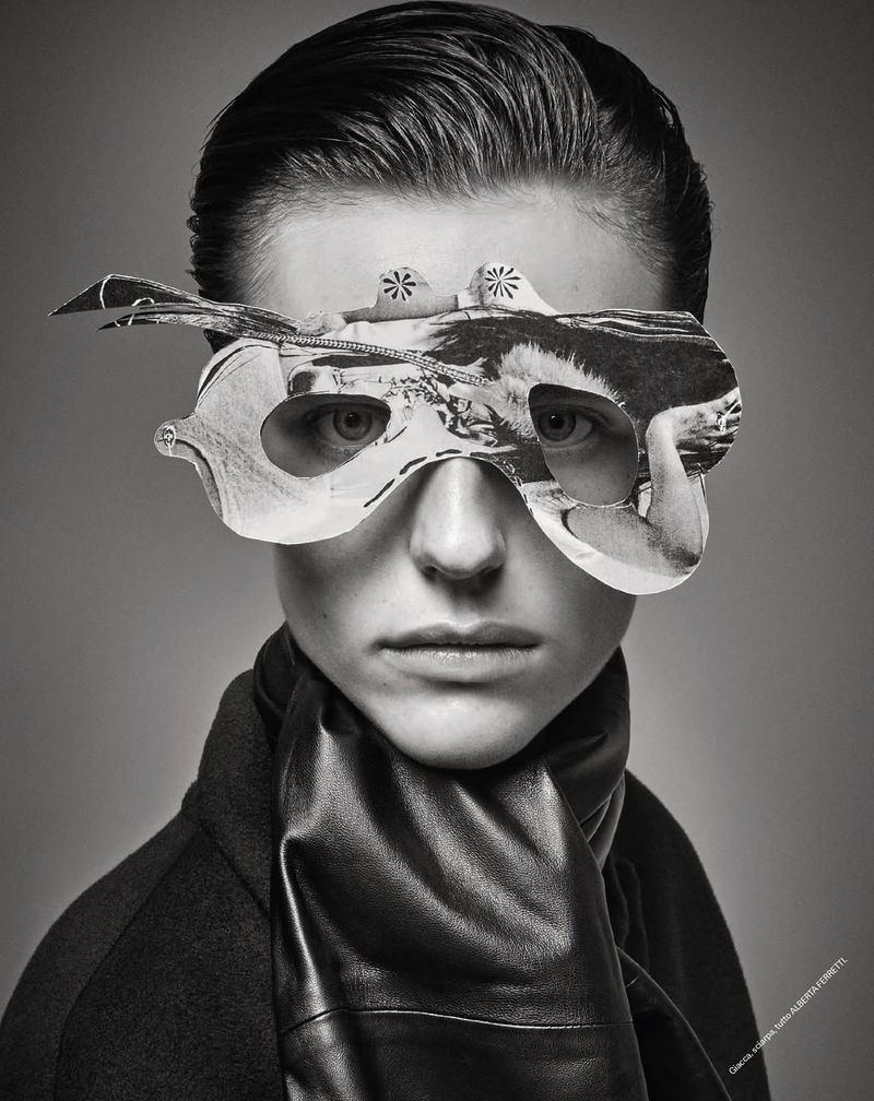 Portrait of a person wearing a unique paper mask and leather scarf against a grey backdrop, showcasing creative and fashion elements.