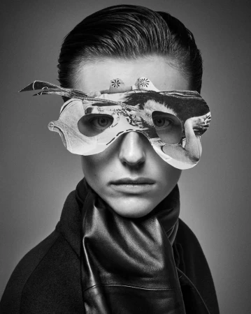 Monochrome portrait of a person with a paper mask over the eyes, wearing a dark coat and leather scarf against a grey backdrop.
