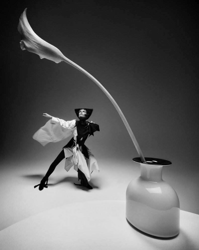 Black and white fashion shot of a woman in an avant-garde outfit beside a white ceramic vase with a calla lily. 