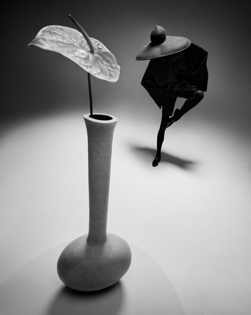 Monochrome studio still life with a white anthurium in a grey vase, a figure in a wide-brimmed hat, and a draped garment.