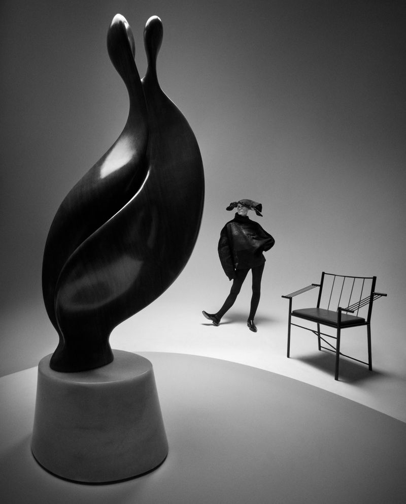 Monochrome studio shot of a modern sculpture alongside a fashion model in black with a chair, creating a stark, stylish composition.