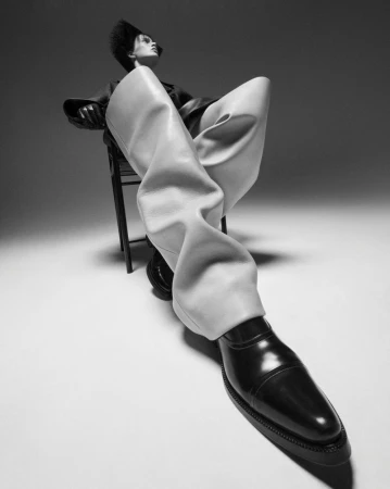 Fashion shot of an adult female model in studio with black top, cream trousers and black shoes sitting on chair with white background.