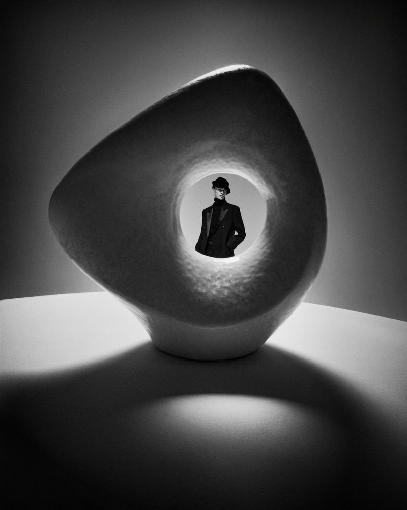Monochrome fashion shot of an adult wearing a hat and suit jacket, viewed through a circular cutout in a sculptural form.