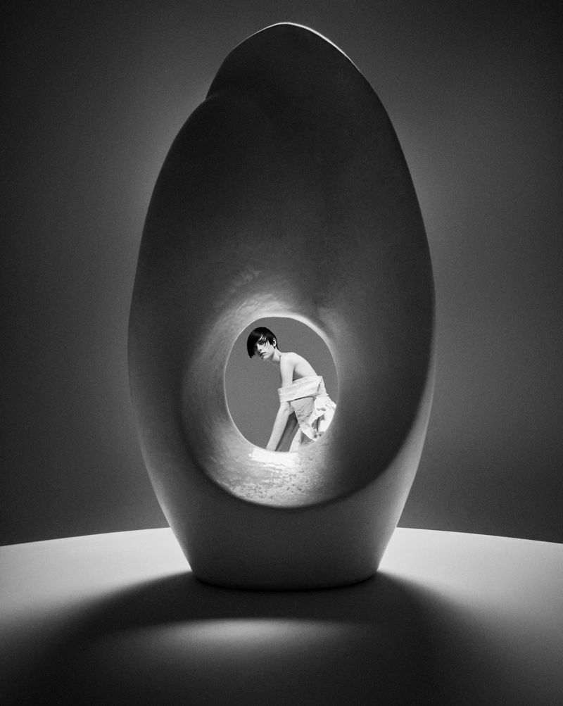 Monochrome photo of a woman standing behind a large decorative sculpture with a hole in the center, posing in a photo studio.