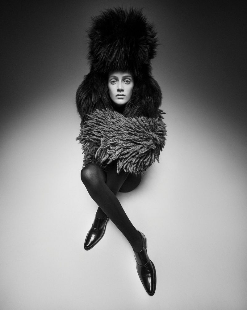Black and white studio shot of a woman wearing a fur hat and coat with legs crossed, in black tights and shoes, against a grey gradient background.