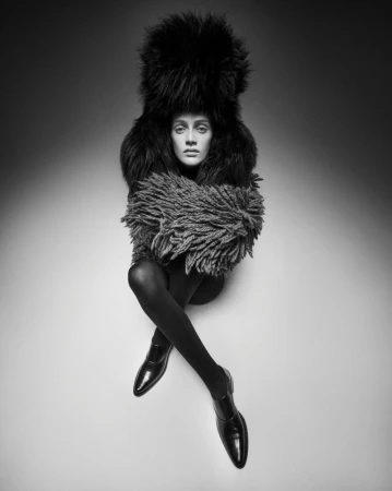 Black and white studio shot of a woman wearing a fur hat and coat with legs crossed, in black tights and shoes, against a grey gradient background.