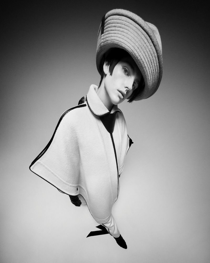 A model in a light-colored cape with dark trim and a wide-brimmed hat, captured in a studio shot with a wide angle lens, in black and white.