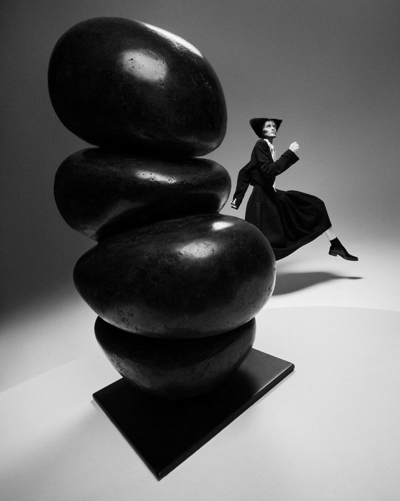 Black and white studio shot of a woman in dark clothing leaping to the right of a sculpture of stacked oval stones.