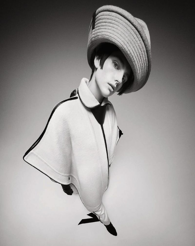 Stylish portrait of a young adult woman wearing a wide-brimmed hat and a fashionable poncho-style coat in a black and white studio shot.
