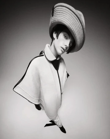 Stylish portrait of a young adult woman wearing a wide-brimmed hat and a fashionable poncho-style coat in a black and white studio shot.