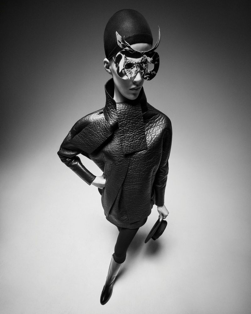 Fashionable woman in a black quilted dress, tights, and boots, posing with a sculptural mask and holding a hat in a studio shot.