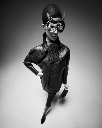 Fashionable woman in a black quilted dress, tights, and boots, posing with a sculptural mask and holding a hat in a studio shot.