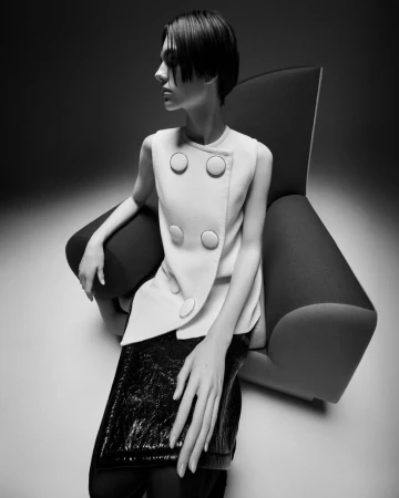 Black and white fashion shot of a slim, fair-skinned woman in a buttoned vest and shiny skirt, posed in an arm chair.