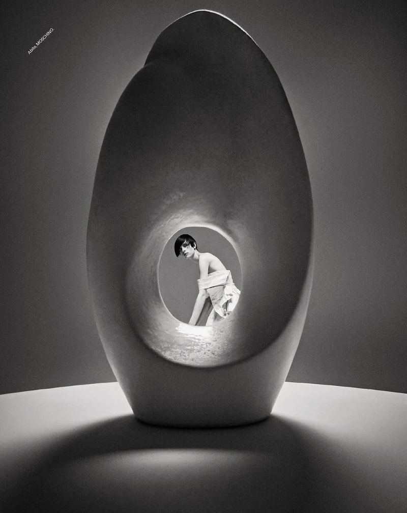 Monochromatic image of a fashion model behind an abstract sculpture, staged indoors with studio lighting and visible shadows.