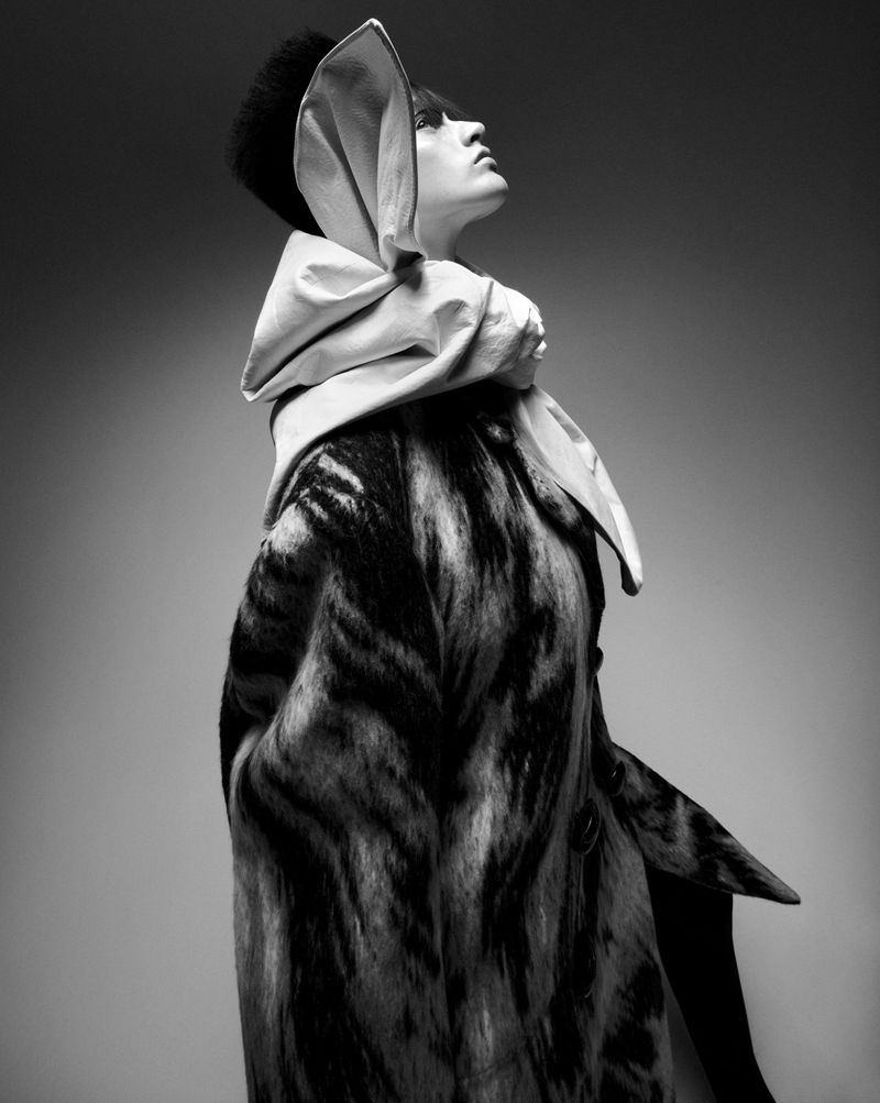Black and white fashion shot of a woman in a patterned coat and large decorative head covering, looking up against a studio backdrop.