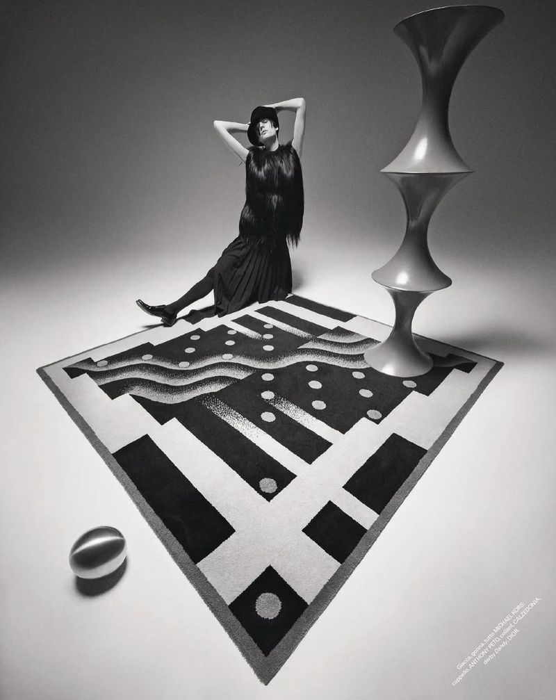 High-angle shot in photo studio of an adult woman in dark textured clothing sitting atop a patterned carpet, with artistic objects.