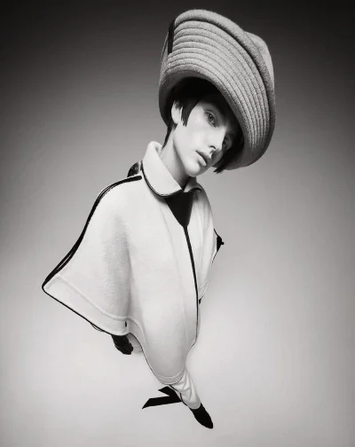 A model wearing a large rounded hat and pale cape coat photographed from a high angle in black and white.