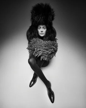 Model seated with legs crossed wearing a massive furry hat and textured coat against a plain gradient background