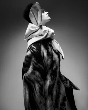 Model in profile wearing a structured white headpiece and a dark patterned coat against a gradient studio background.