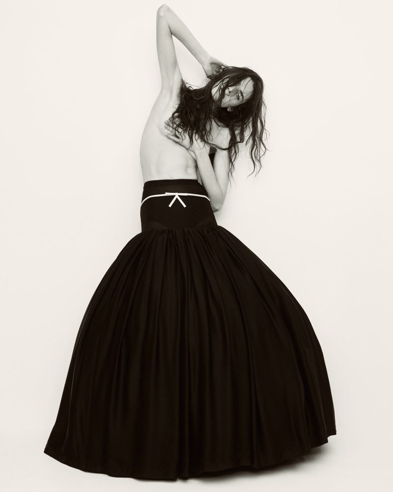 Fashion shot of a woman in a black skirt against a white backdrop, posing with arms overhead and a neutral expression.