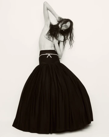 Fashion shot of a woman in a black skirt against a white backdrop, posing with arms overhead and a neutral expression.