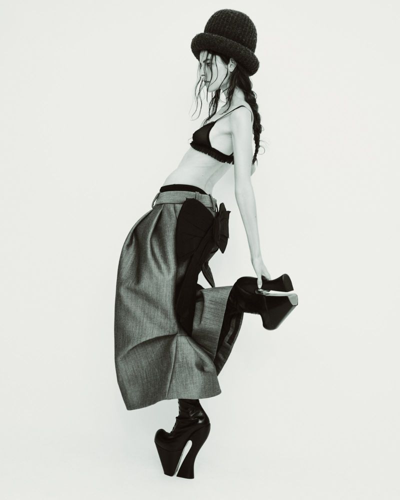 Studio fashion shot of a posing female model in a bra top, high-waisted skirt, hat, and platform heels against a white backdrop.
