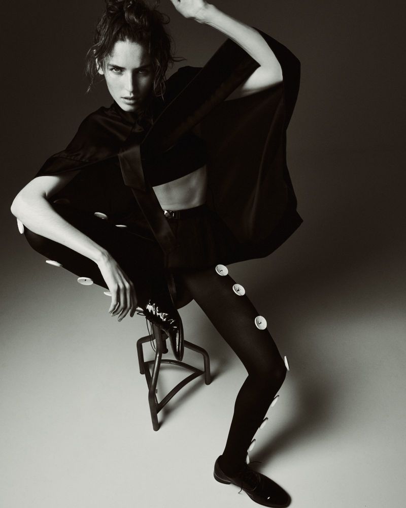 A stylish adult female poses dynamically indoors in a black top, pants with buttons, and dress shoes on a stool against a neutral background.
