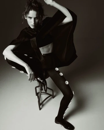 A stylish adult female poses dynamically indoors in a black top, pants with buttons, and dress shoes on a stool against a neutral background.