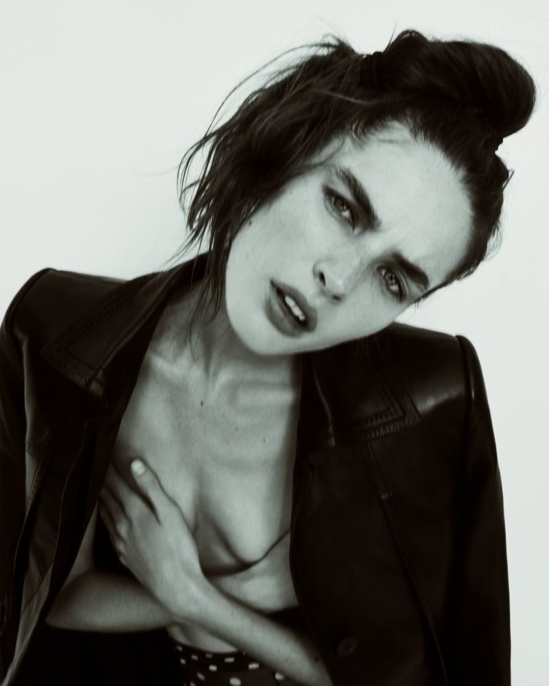 Model Libby Taverner posing with tilted head wearing an open dark leather jacket against a light backdrop.