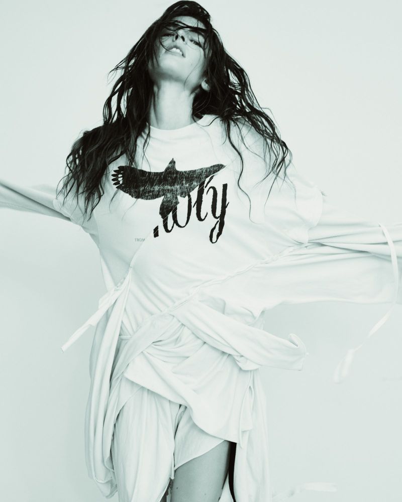 Woman with long dark hair poses in a studio wearing a white long-sleeved shirt and shorts set, with a graphic print on the shirt.