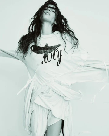 Woman with long dark hair poses in a studio wearing a white long-sleeved shirt and shorts set, with a graphic print on the shirt.