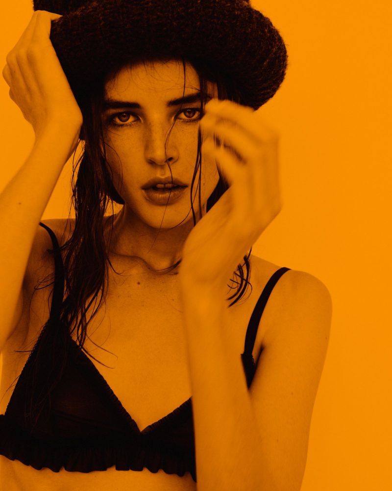 Portrait of a brunette adult female model in a black bra and knit hat, looking at the camera with hands near her face on orange background.