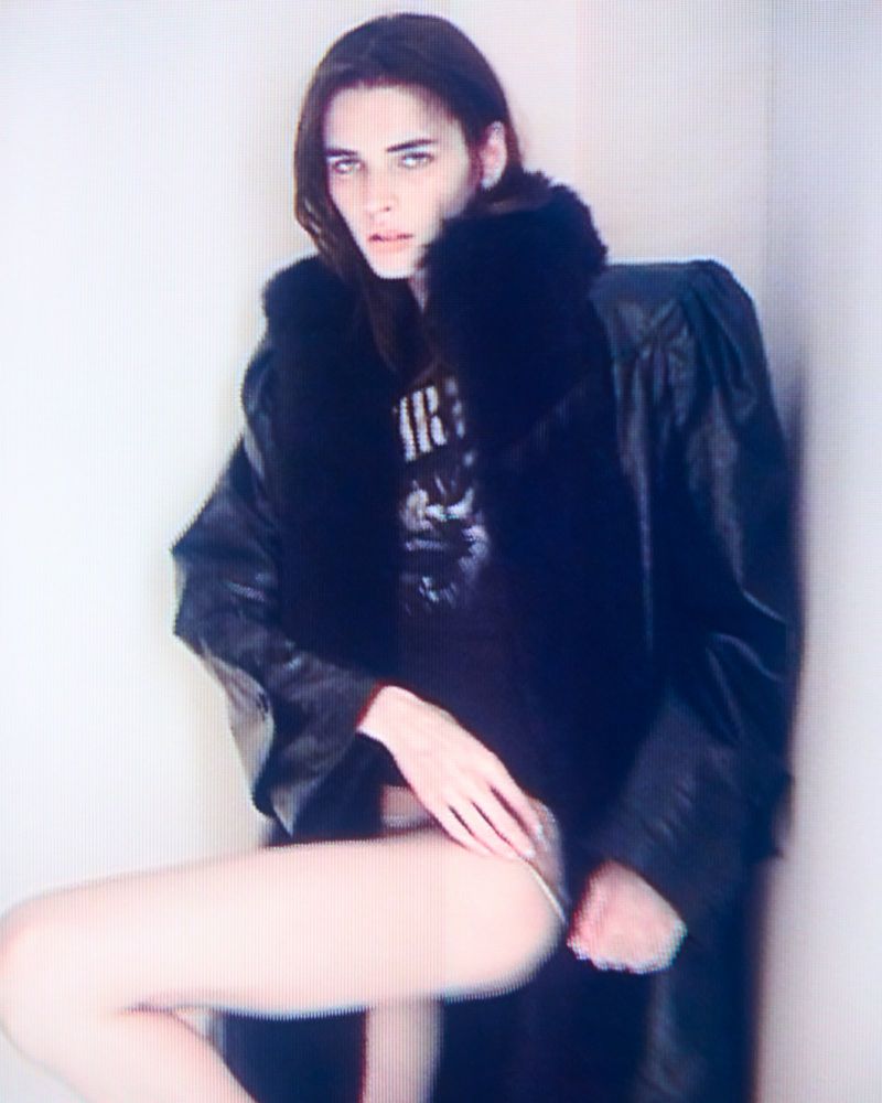Woman in black coat poses indoors. Close-up shot highlights long hair, fair skin, and fashion apparel against a neutral backdrop.