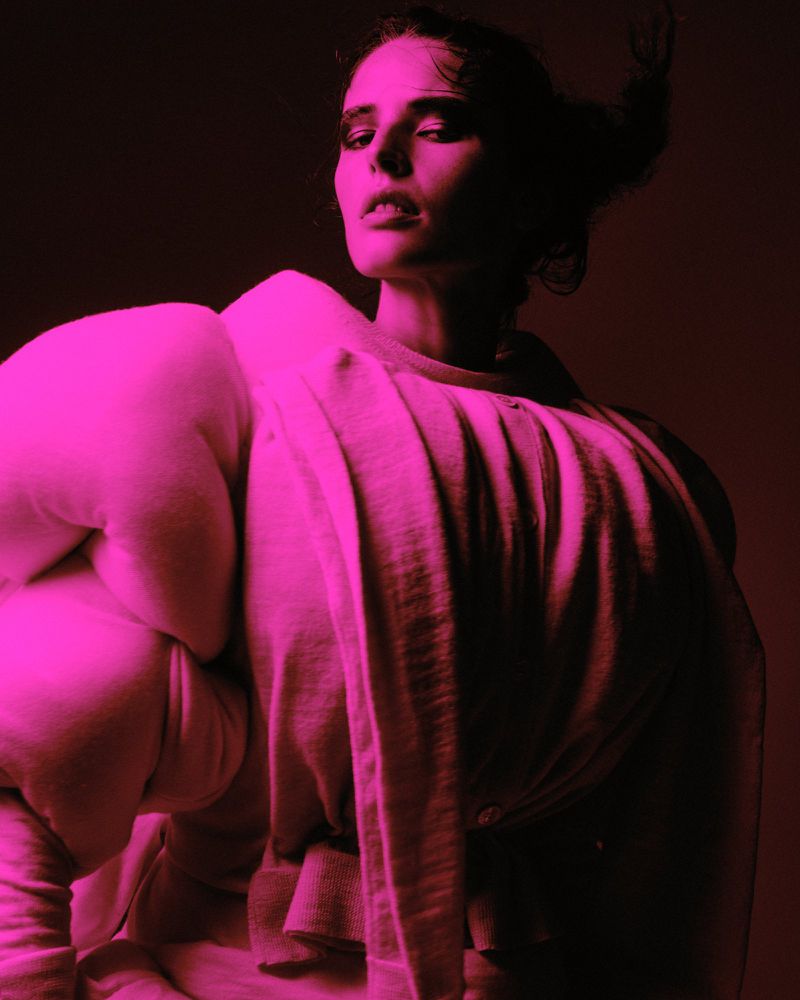 Portrait of an adult female model wearing a pale sweater with exaggerated shoulders, against a dark background with pink lighting.