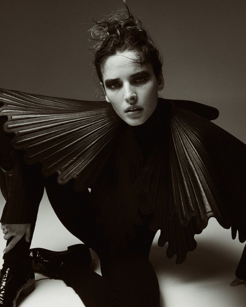 Monochrome fashion shot of a woman kneeling in a dark outfit with a large ruffled collar against a neutral gradient background.