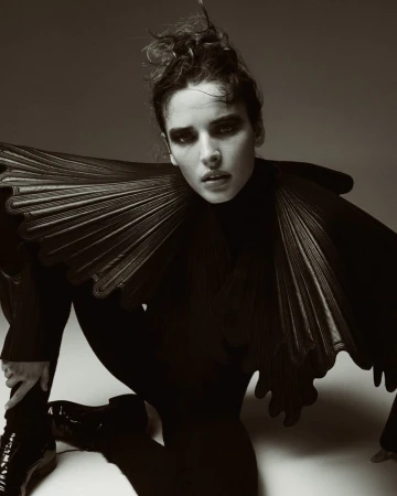 Monochrome fashion shot of a woman kneeling in a dark outfit with a large ruffled collar against a neutral gradient background.