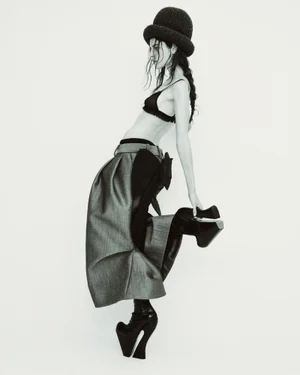 Model Libby Taverner wearing a rounded hat, black bra top, sculptural grey trousers, and curved platform boots against a white backdrop.