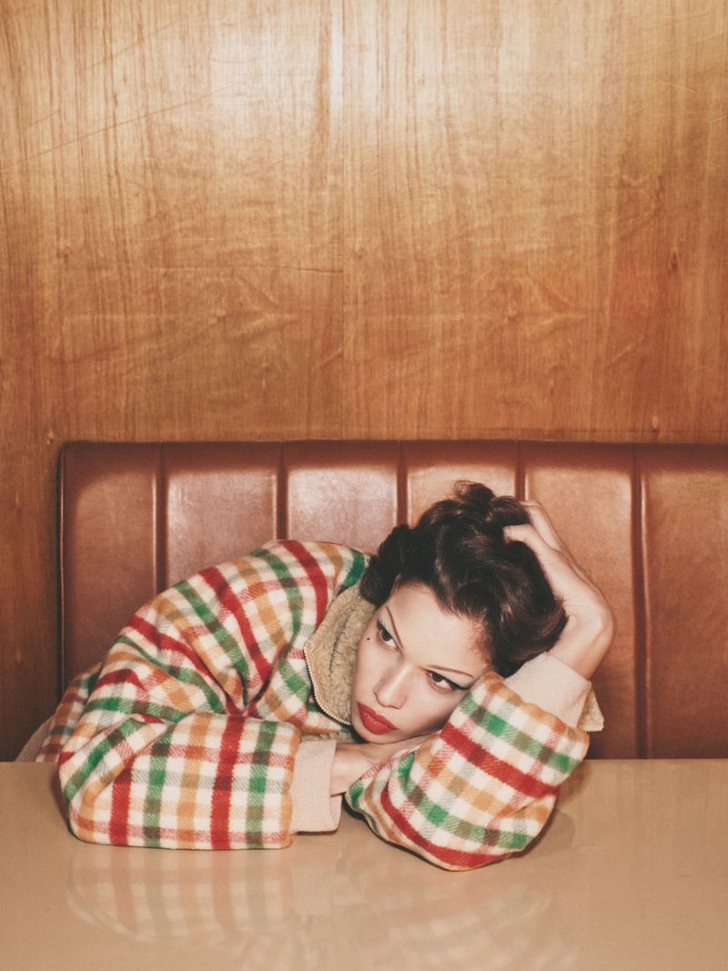 Indoor portrait of an adult female with her head resting on a beige table in a plaid top, seated against a wood-paneled wall.
