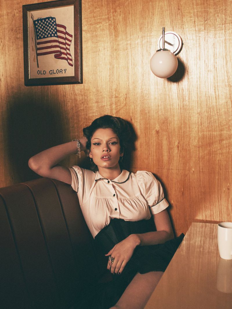 Woman in vintage-style blouse poses in diner booth. Wood paneled wall, framed US flag art, and table lamp add to the retro scene.