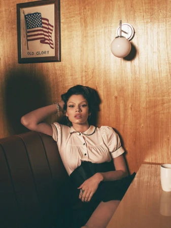Woman in vintage-style blouse poses in diner booth. Wood paneled wall, framed US flag art, and table lamp add to the retro scene.