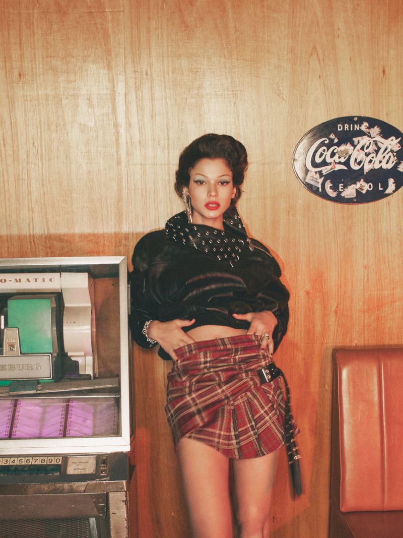 Woman in a plaid skirt and black jacket poses next to a vintage jukebox and Coca-Cola sign in a retro-styled indoor setting.