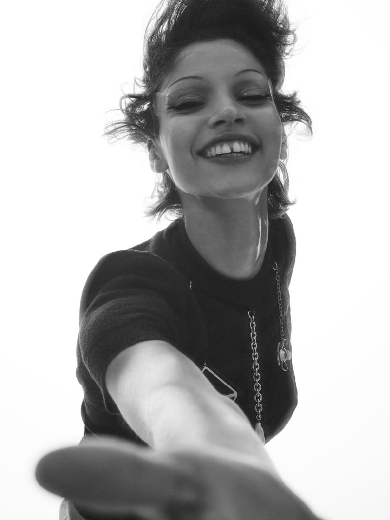 Black and white portrait of an adult female with short dark hair reaching out with her hand, smiling, wearing a dark top.