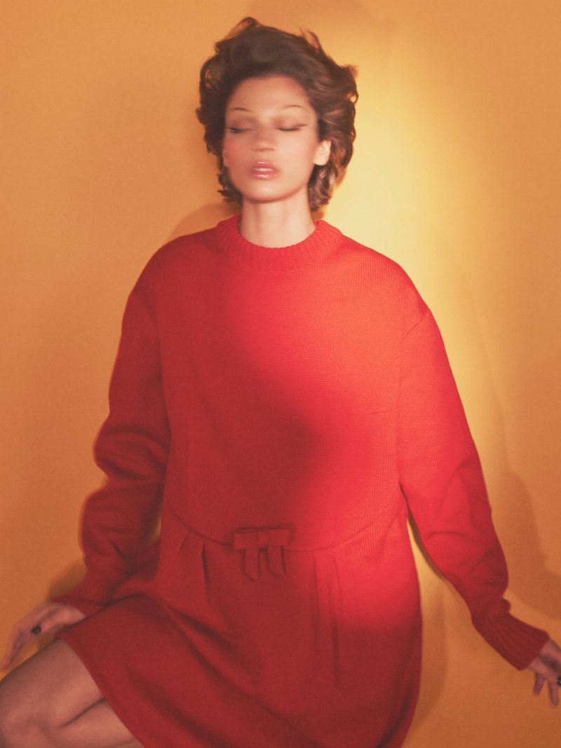 Young woman with short brown hair wearing a red dress, lit by warm light in a photo studio with an orange-toned background.
