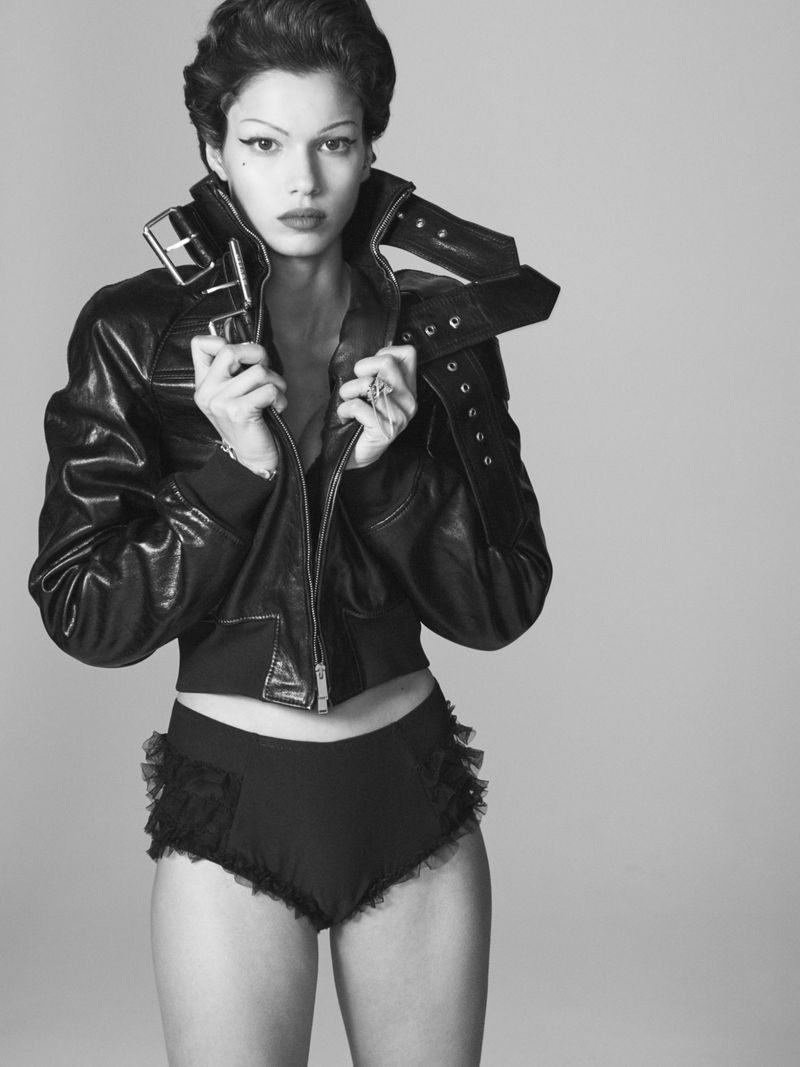 Black-and-white portrait of a woman in a leather jacket and ruffled shorts posing in a studio with short hair and makeup.