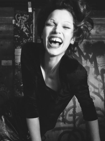 Close-up black and white shot of a young woman with open mouth laughing, wearing a dark long-sleeve shirt with a graffiti background.