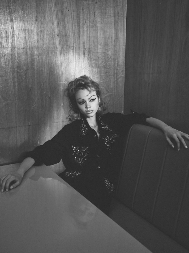 Monochrome portrait of a woman with styled hair and a decorative shirt sitting in a booth indoors, captured with natural lighting.