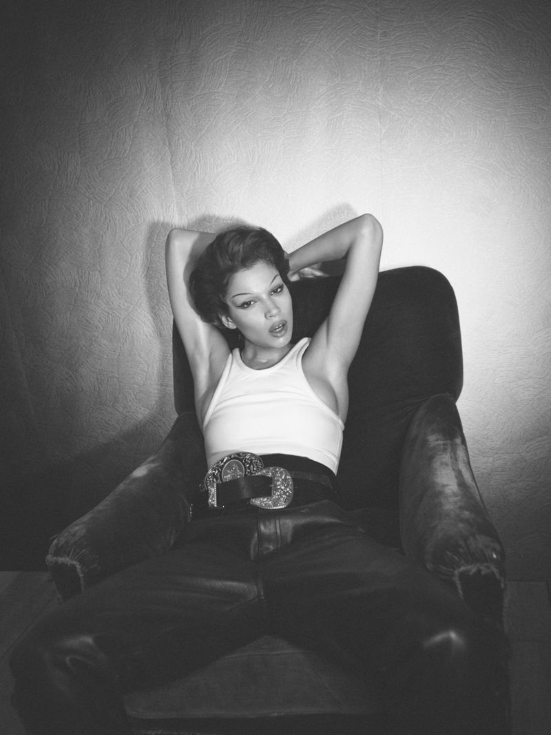 Model seated in an armchair with arms behind head, wearing a white cropped tank top and dark leather trousers with an ornate belt buckle.