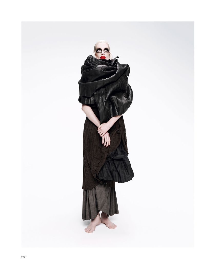 Woman stands in a studio, wearing avant-garde black clothing with red lipstick and heavy eye makeup, against a white backdrop.