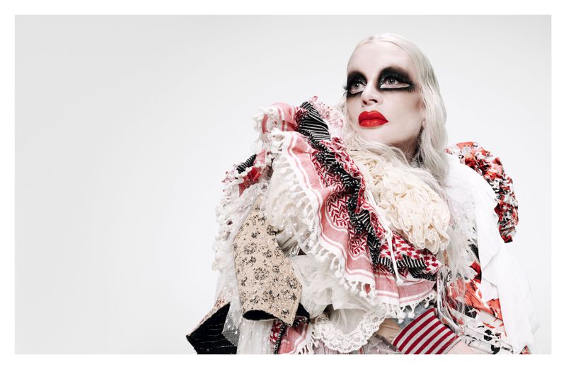 Artistic portrait of a woman with heavy makeup and a theatrical costume with various patterns and textures, looking up against a white backdrop.