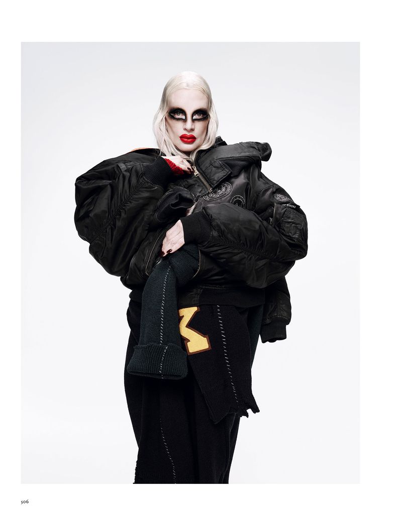 A person with pale skin and dark makeup, wearing an oversized black bomber jacket and black clothing, poses in front of a white background.